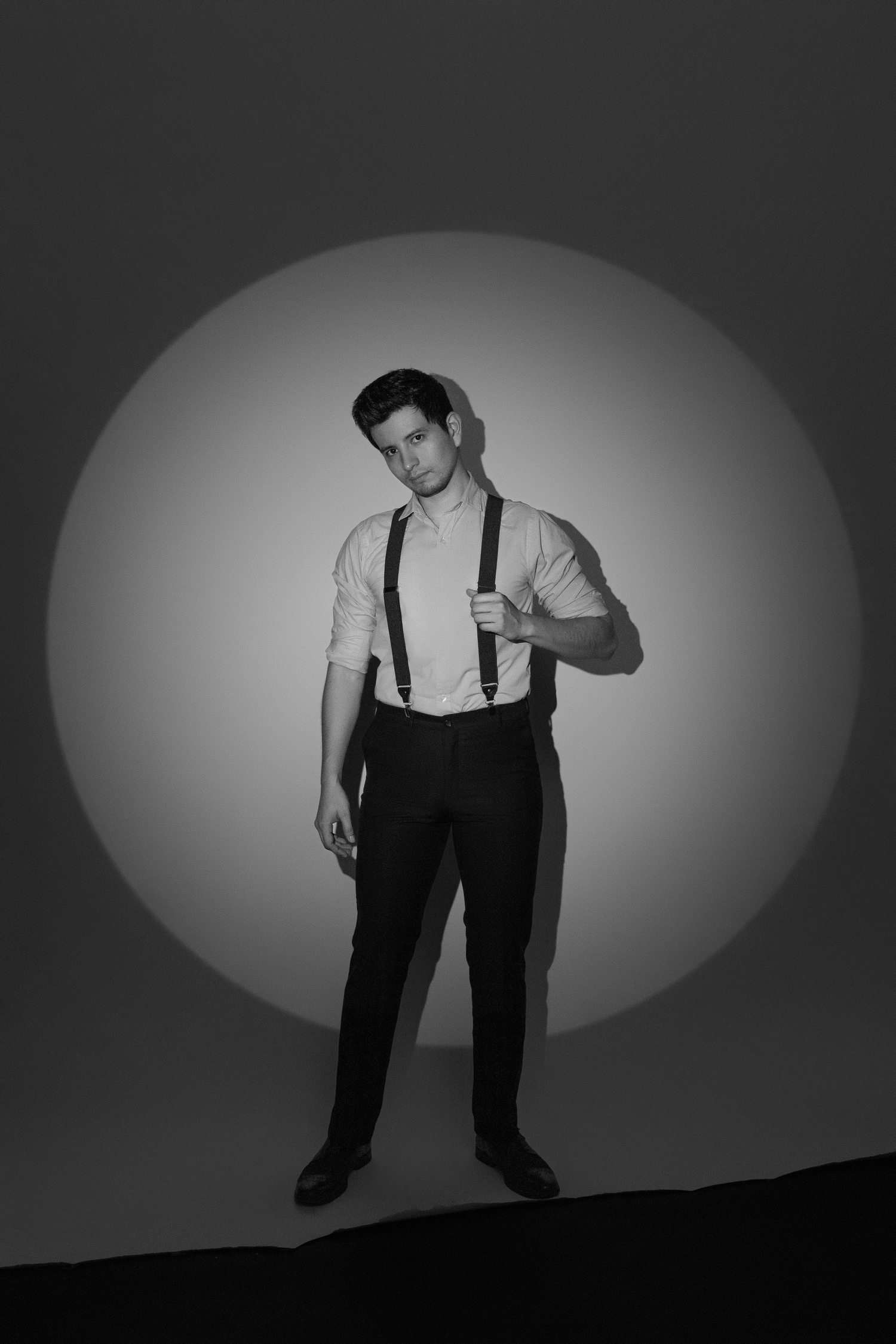 Black and white portrait of a figure wearing suspenders and a white shirt posing against a spotlit circular backdrop.