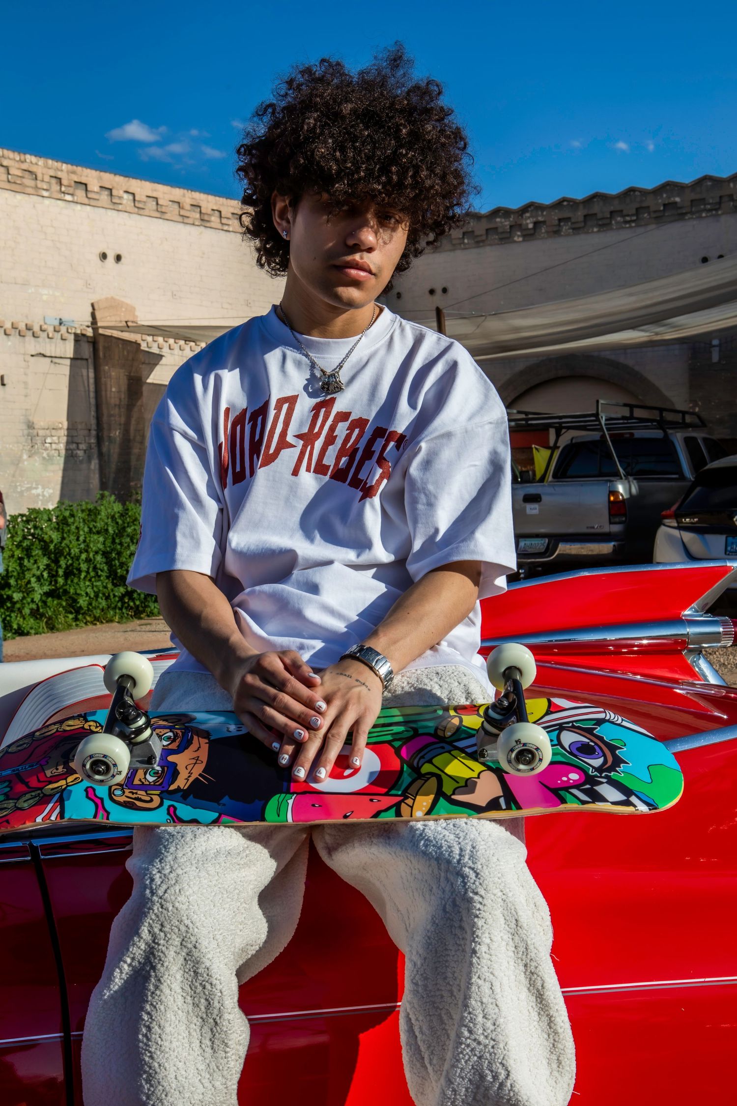 Person in white t-shirt sits on red car hood with colorful skateboard and white sneakers.