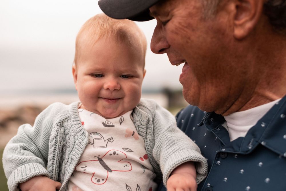 Close up of joyful baby in grey sweater with parent wearing baseball cap.