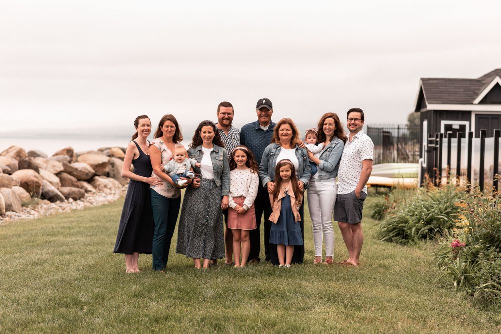 Extended family gathering outdoors with coastal house and rocky shoreline in background.