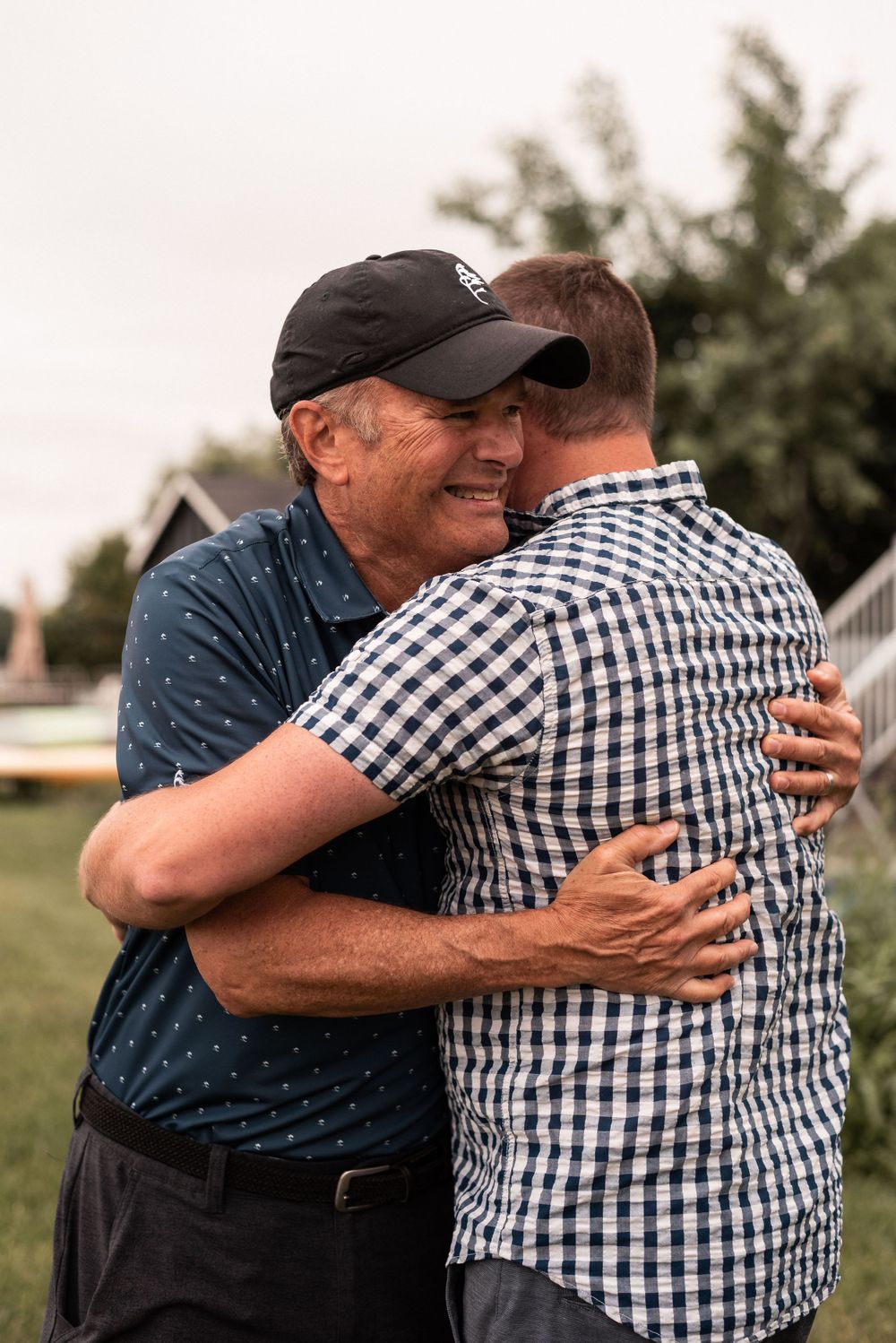 Two people sharing emotional embrace outdoors, one wearing baseball cap and polka dot shirt.