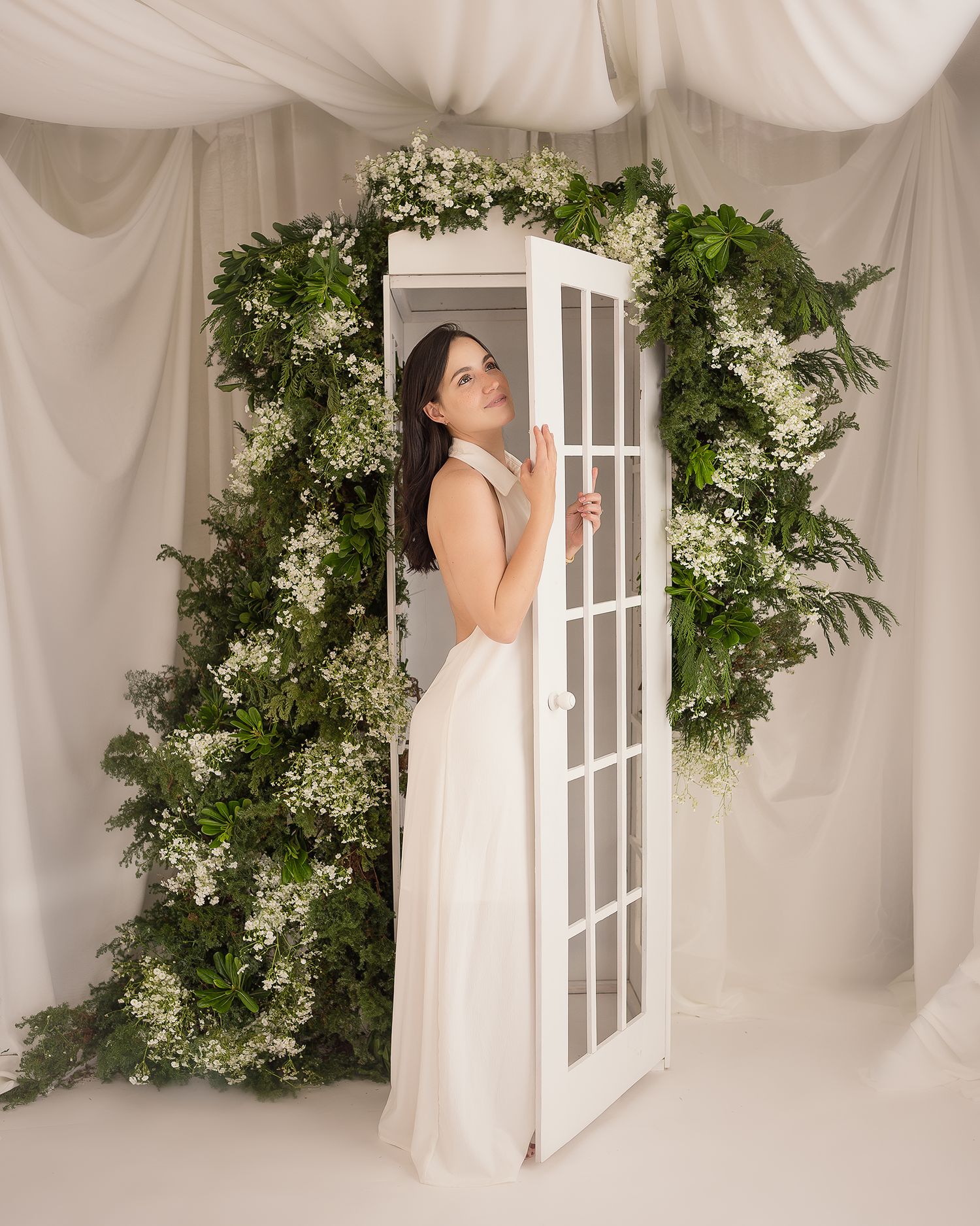 A decorative white door frame surrounded by lush greenery and white floral arrangements against a light fabric backdrop.