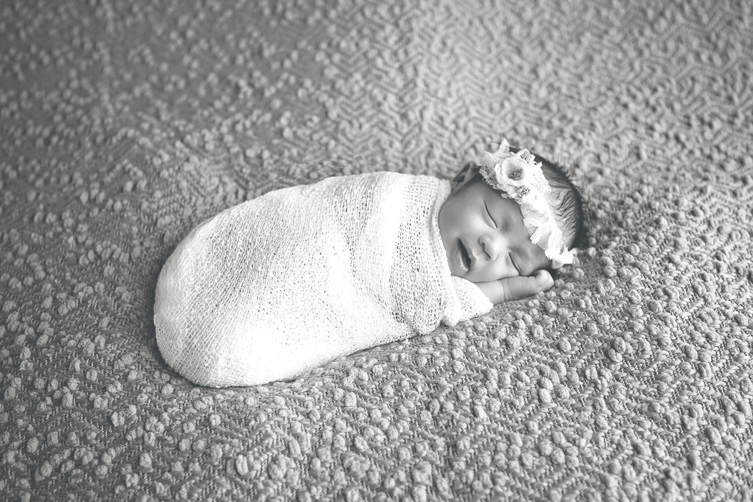 Sleeping newborn baby wrapped in white swaddle on textured gray blanket.