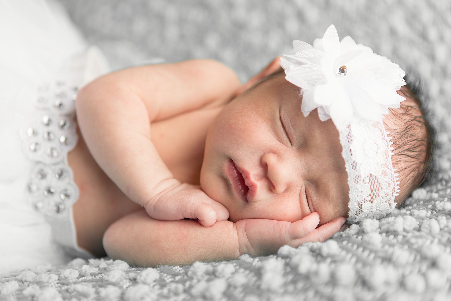 Sleeping newborn wearing delicate white lace headband on textured gray blanket.