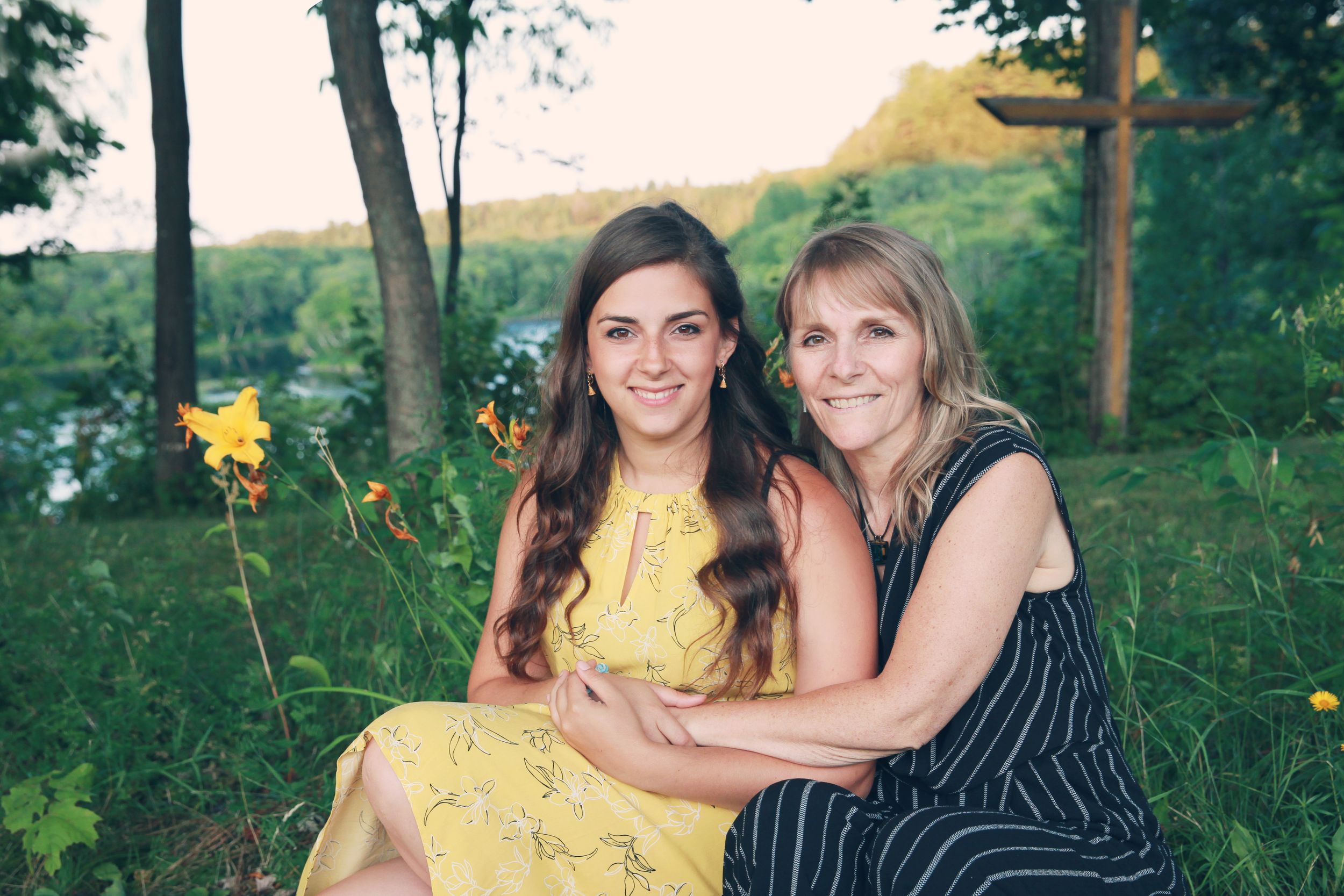 Two women sitting outdoors in summer attire with trees and sunset in background.