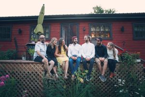 Group of people sitting on wooden deck railing with red building and garden in background.