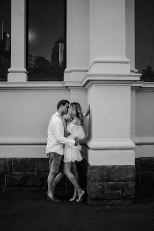 An engaged couple leaning against pillar kissing.