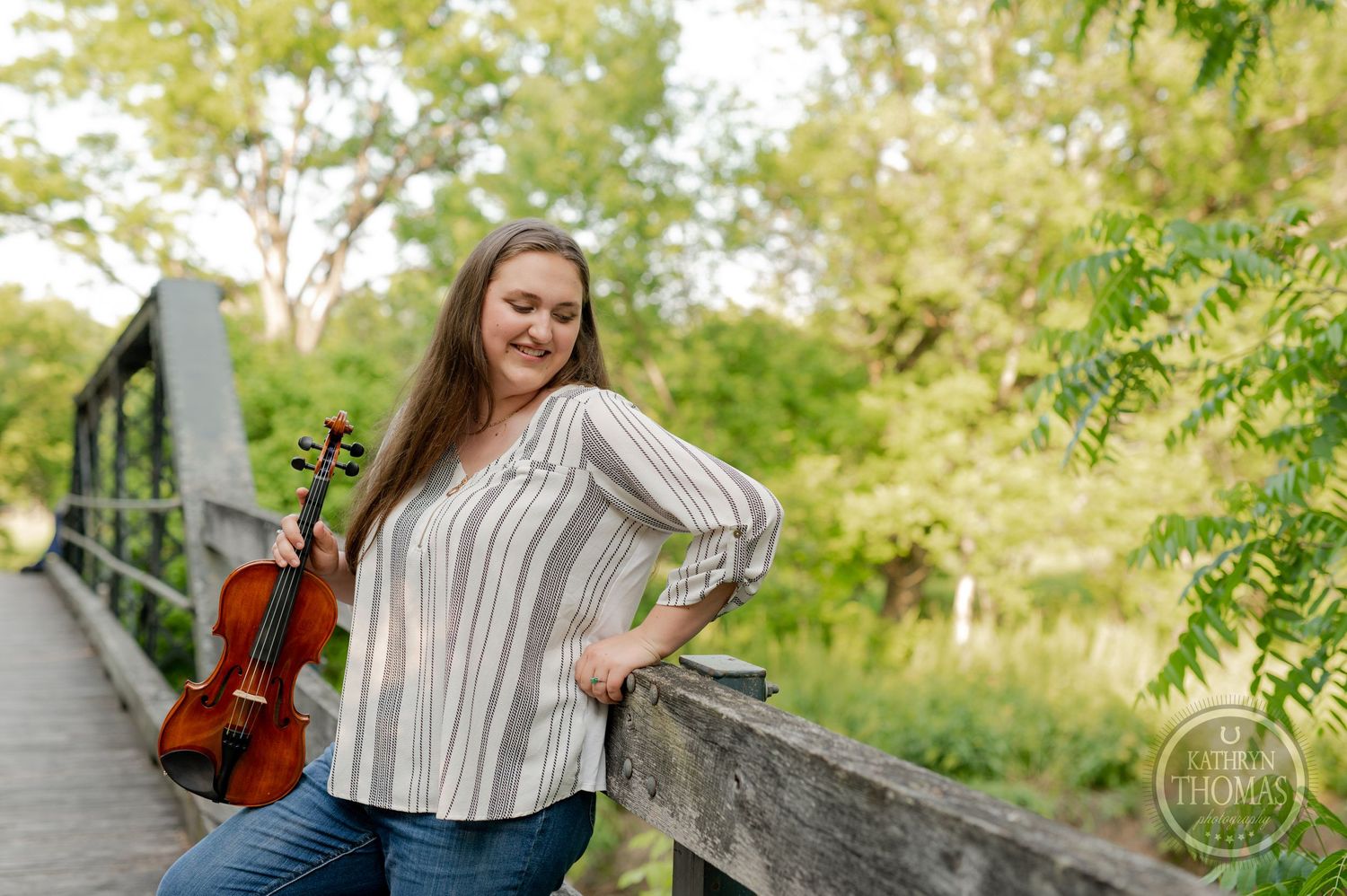 Musician in striped shirt poses with violin on rustic bridge in nature setting.