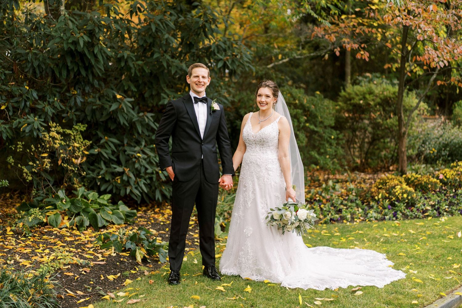 Wedding couple poses together on lawn with fall foliage in background.