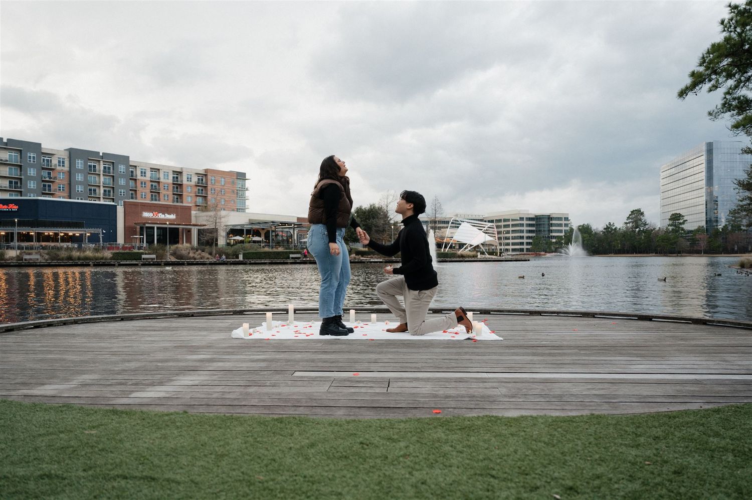 A couple shares a special moment on a waterfront dock overlooking modern buildings and calm waters.