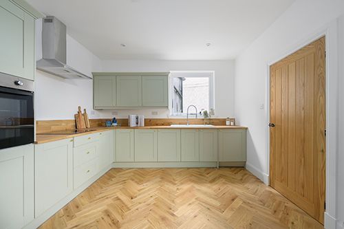 Modern kitchen interior with sage green cabinets, wooden accents, and herringbone hardwood flooring. HMO Photography
