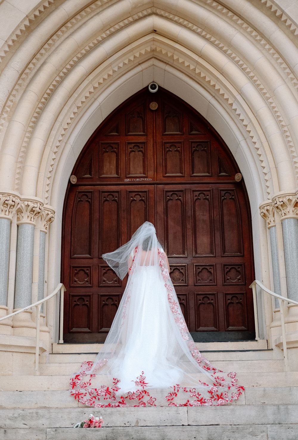 Wedding dress with red floral veil detail against gothic church door architecture.