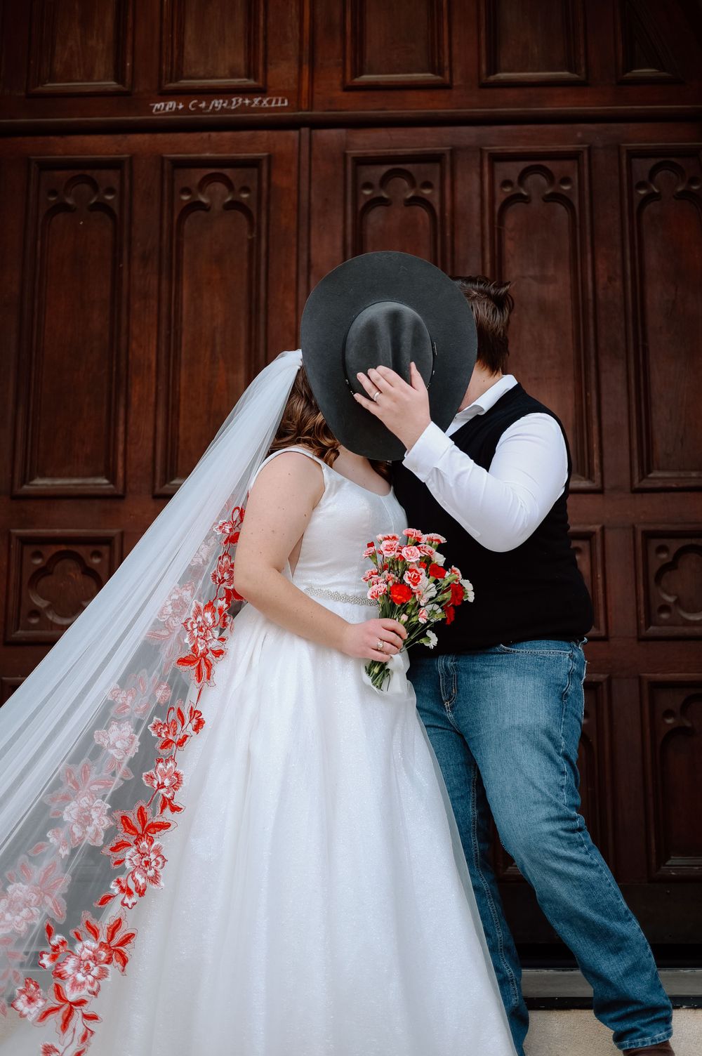 Romantic wedding couple sharing a kiss behind a black hat against wooden doors.