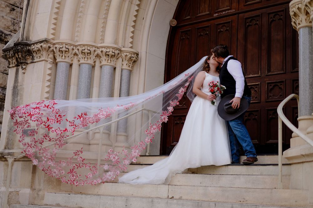 A wedding couple stands in front of ornate church doors with a flowing pink-embroidered veil caught in the breeze.