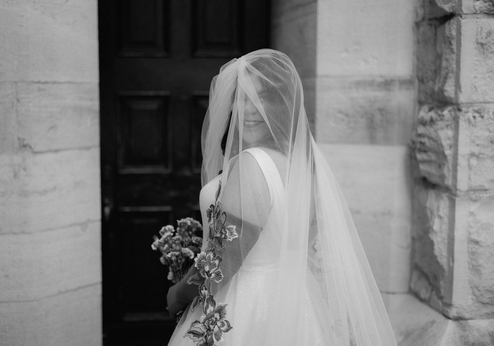 A black and white portrait of a bride with flowing veil standing beside a stone church entrance.
