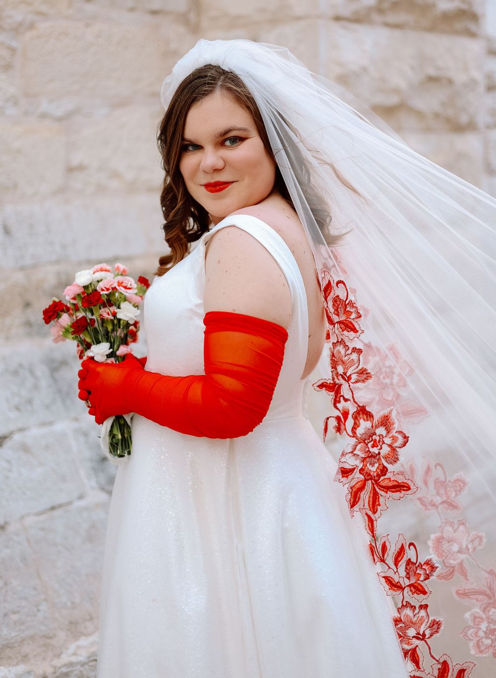 A bride in a white wedding dress with bright red arm sleeves holds a bouquet against a stone wall.