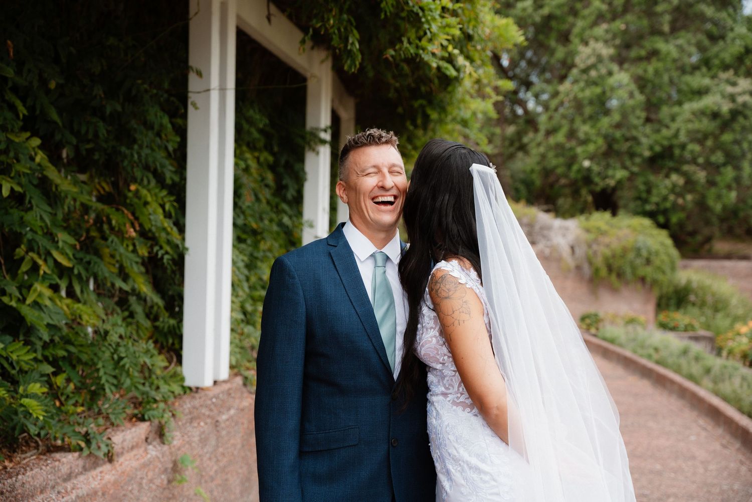 A laughing groom in a navy suit stands with a bride in white dress and veil outdoors at a garden venue.