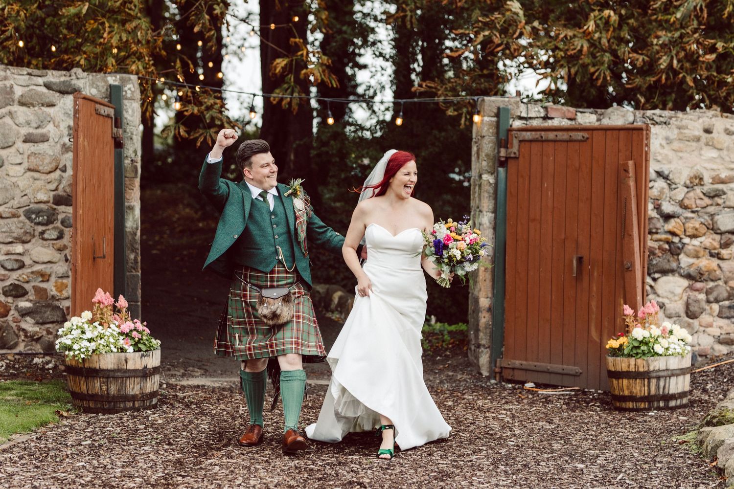 A wedding couple exits through wooden gates with stone walls and barrel planters in a rustic outdoor setting.