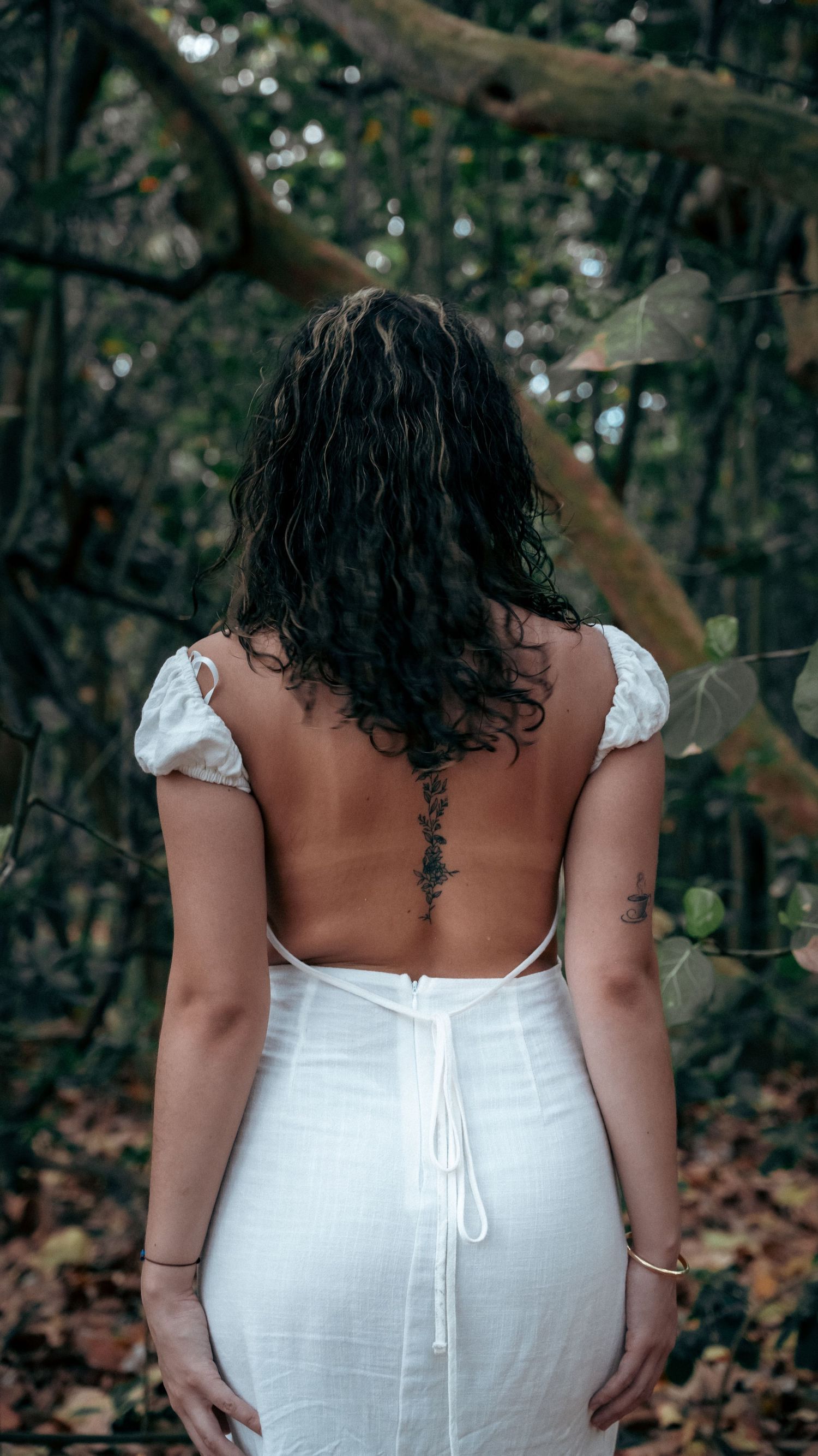 Back view of someone in a white backless dress with dark curly hair in a forest setting.