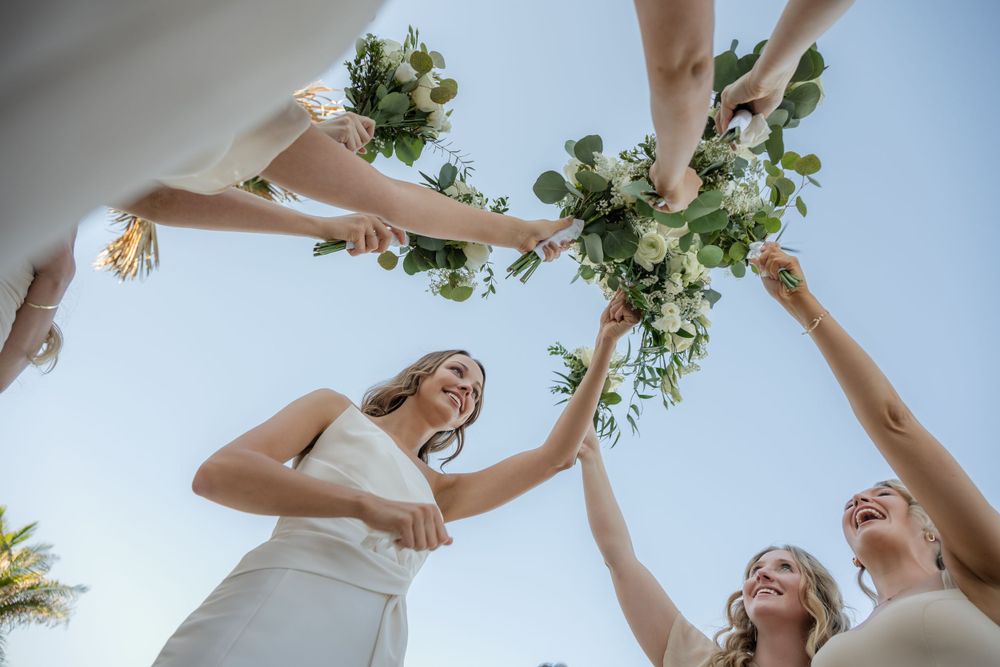 Bridesmaids in light colored dresses raise their bouquets in a celebratory circle against a bright sky.