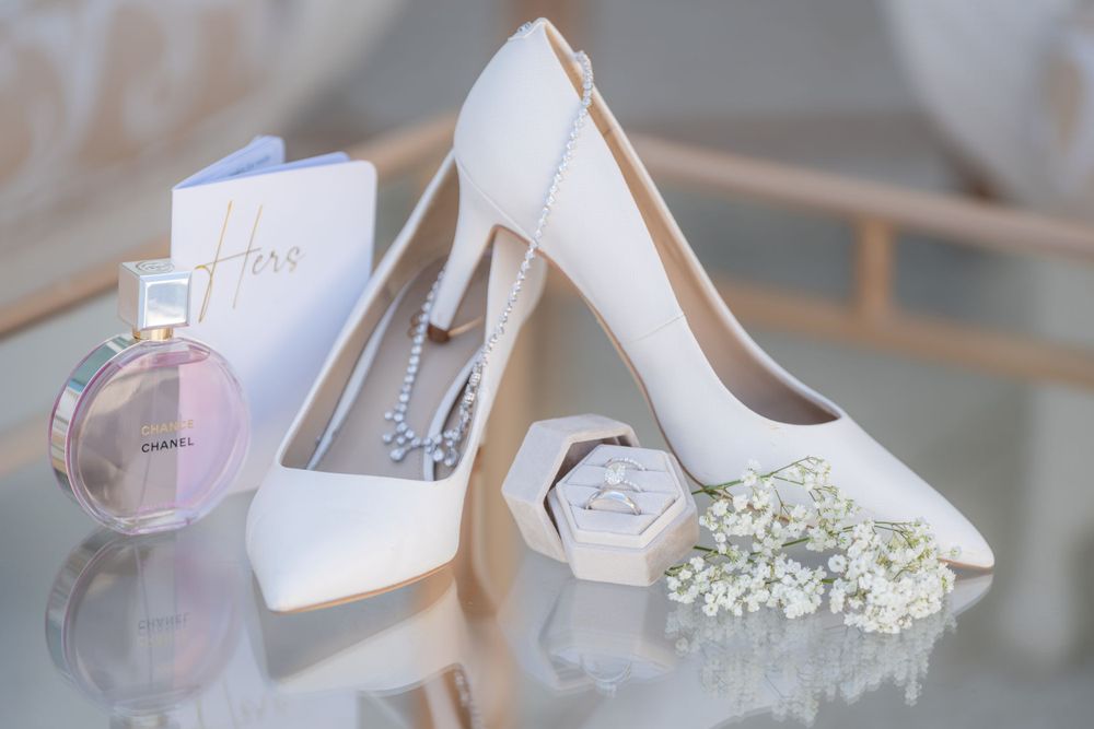 White bridal heels with perfume bottle and baby's breath flowers on reflective surface.