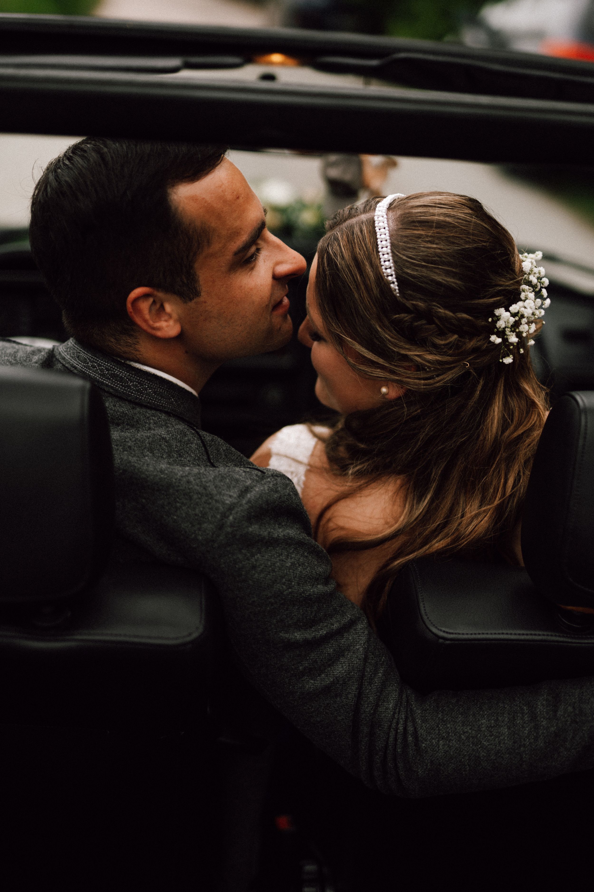 Wedding couple embraces near a vintage car, with the bride wearing a delicate hair accessory.