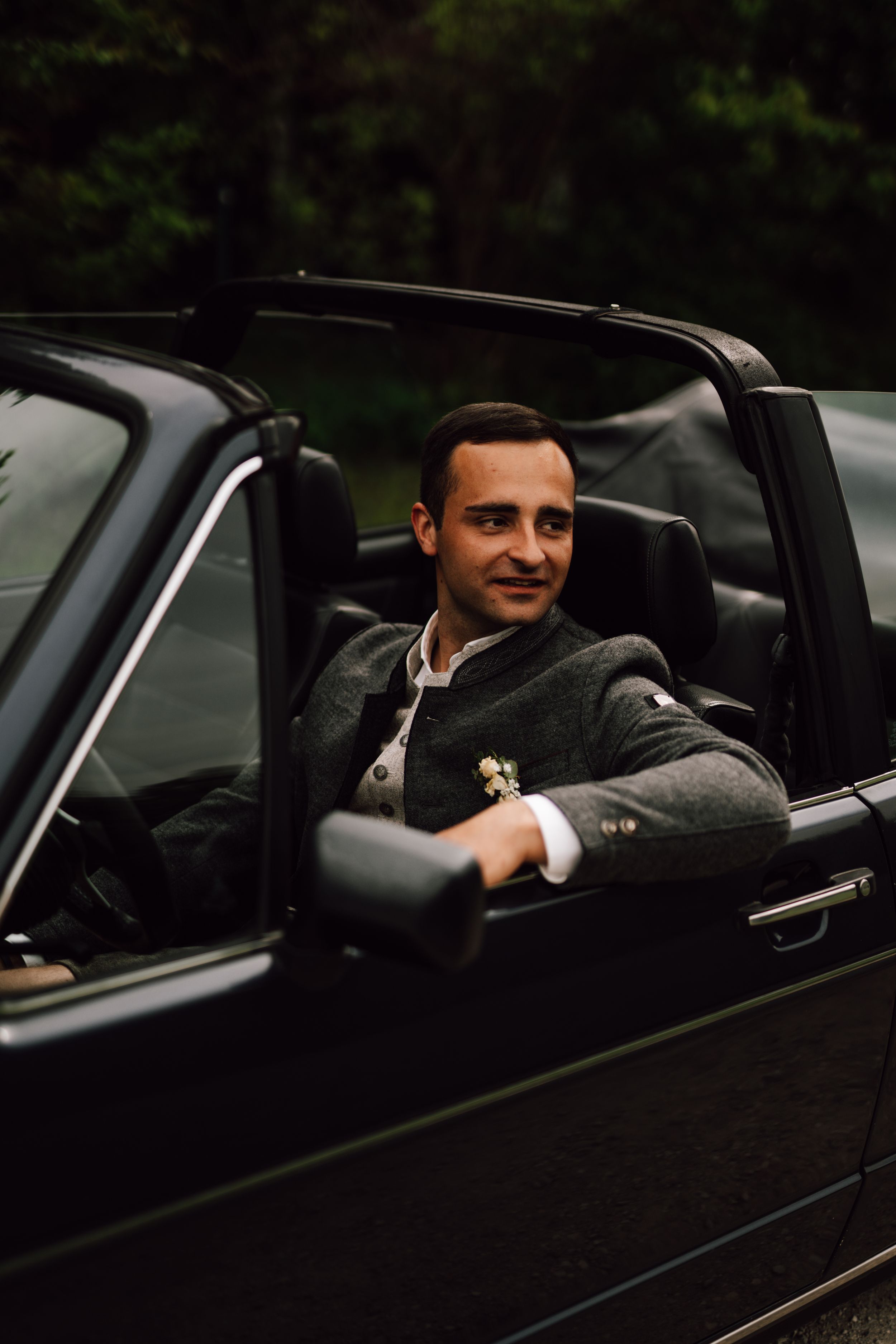 Groom in grey suit sitting in classic black convertible car.