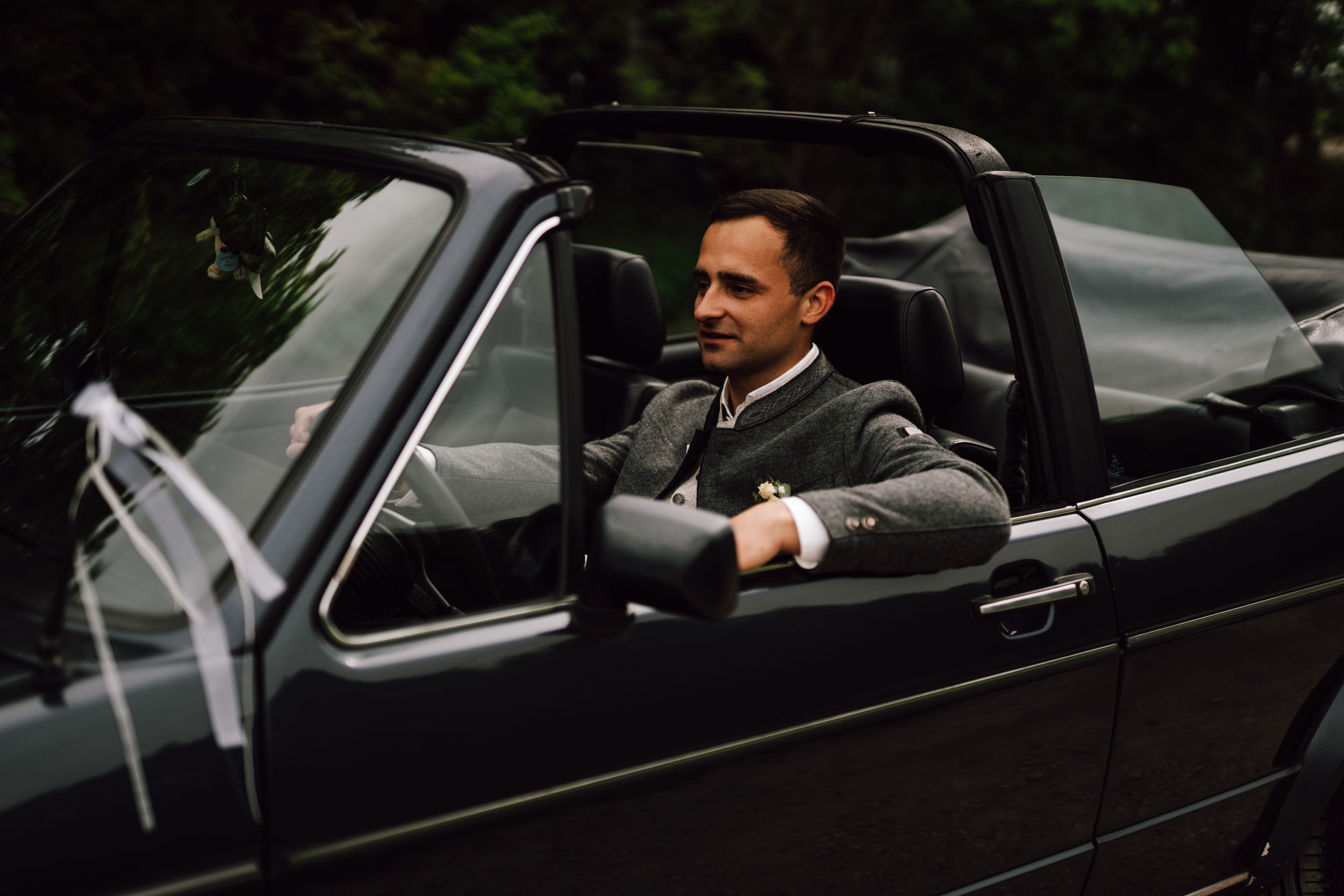 Groom seated in vintage black convertible car wearing grey suit.