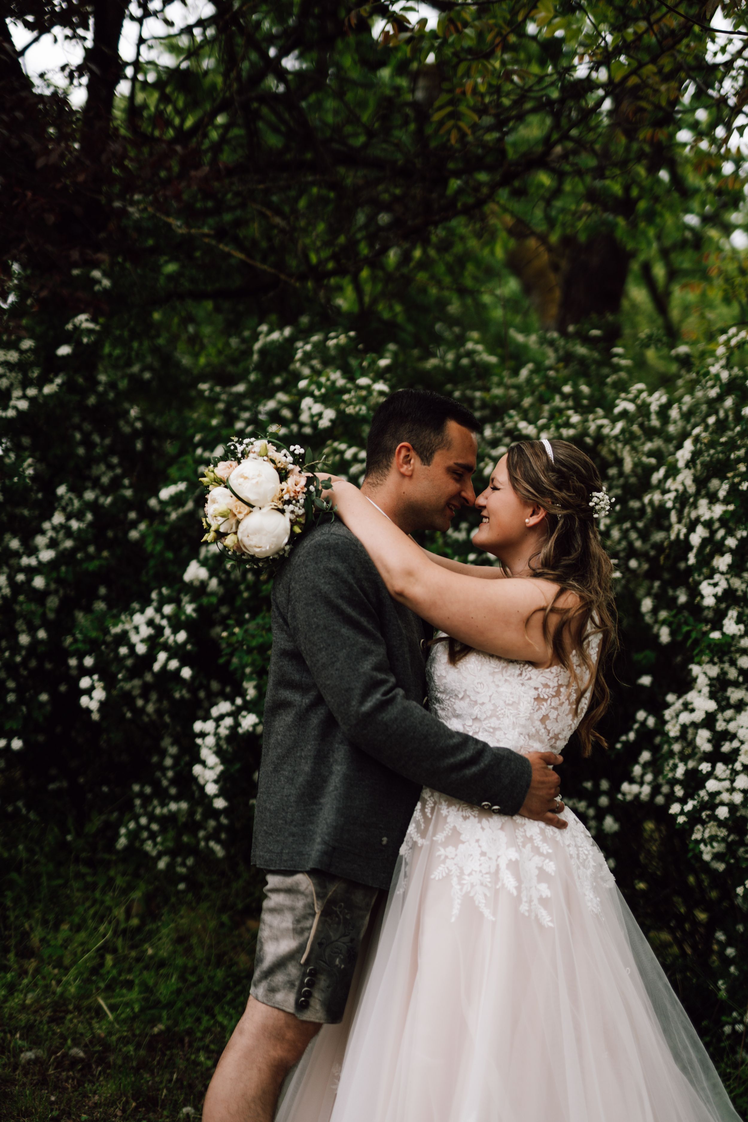 Newlyweds share an intimate embrace surrounded by blooming white flowers and lush foliage.