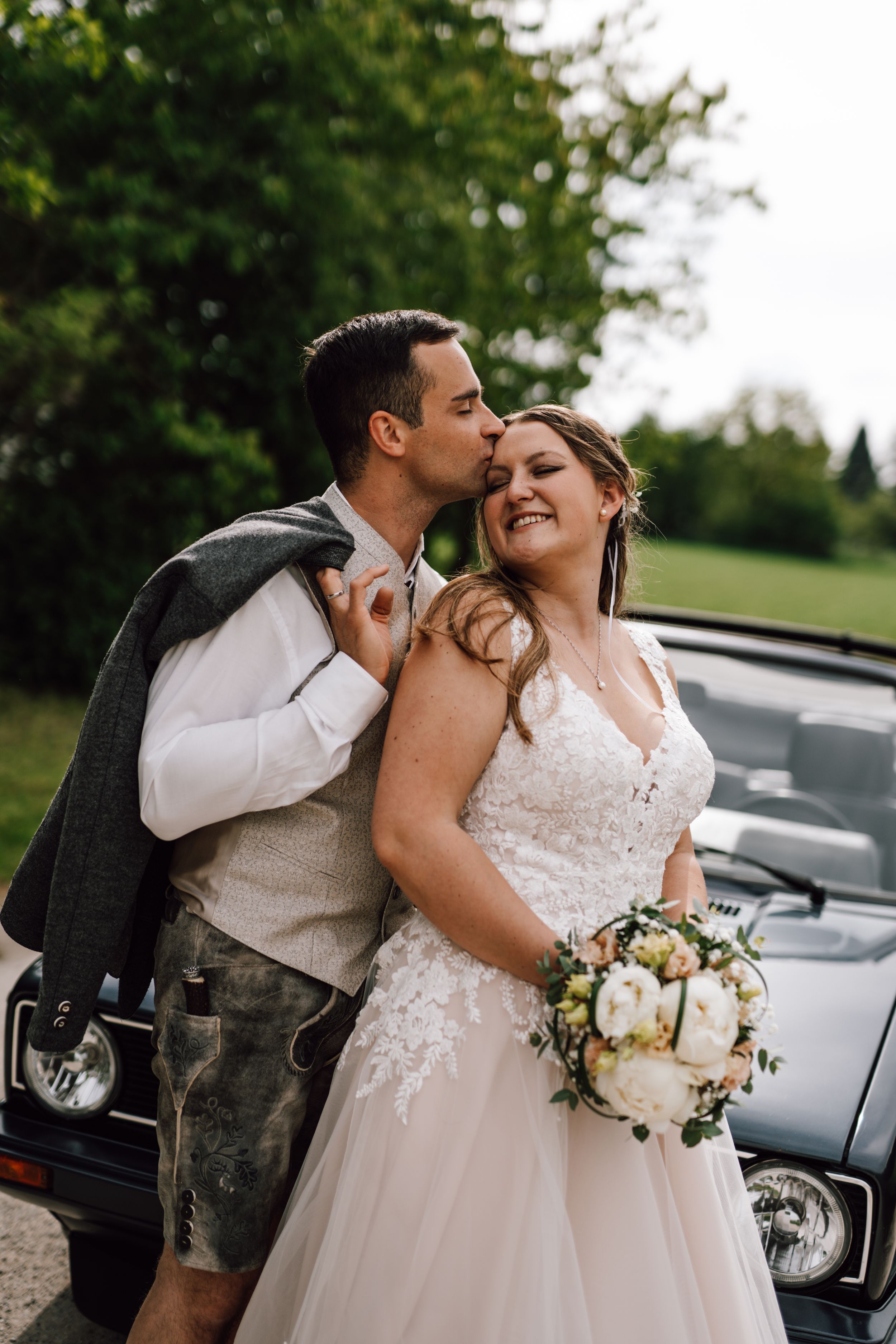 Newlyweds share joyful laughter next to a classic car during their wedding photoshoot.