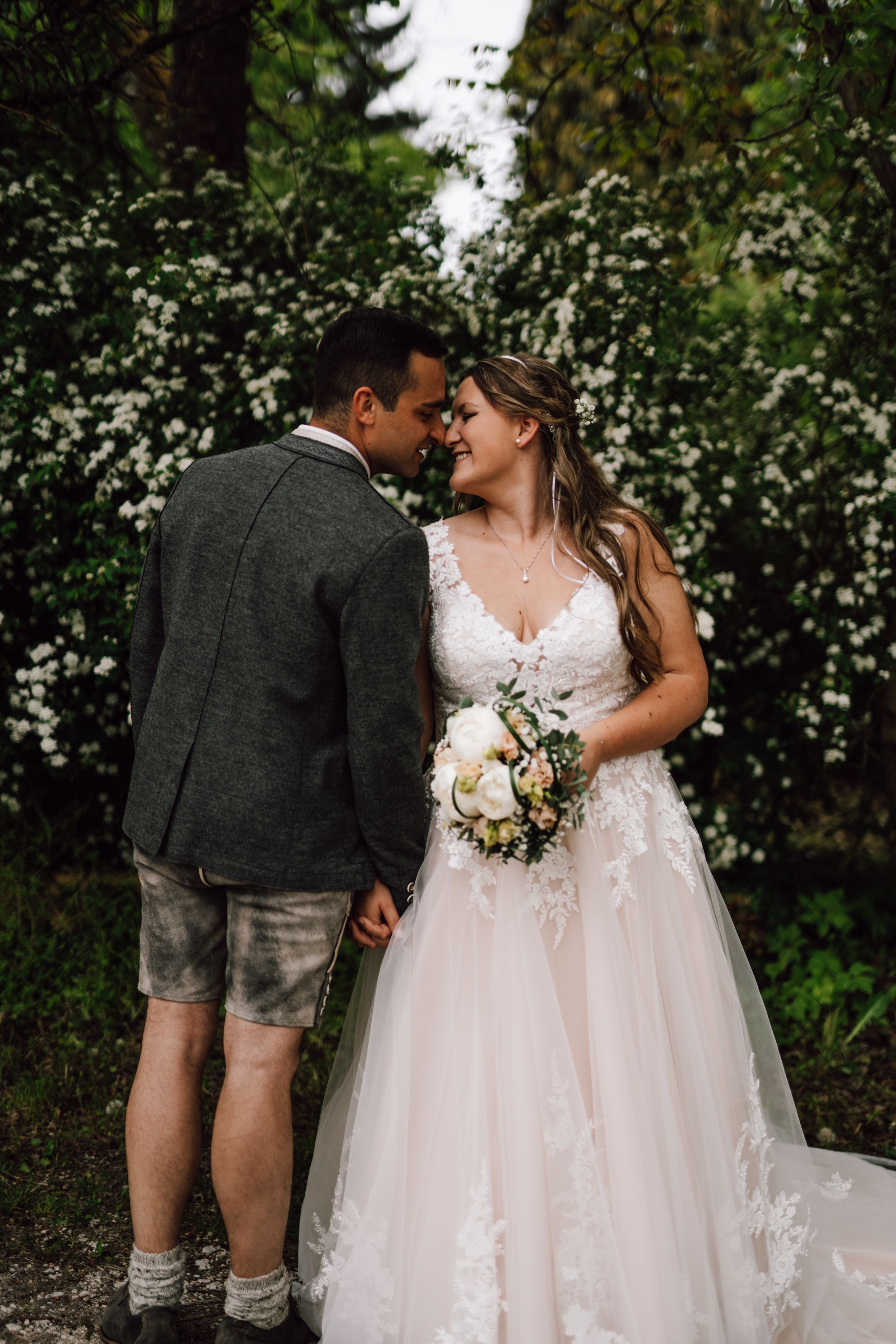 A wedding couple poses together against a backdrop of white flowering bushes.