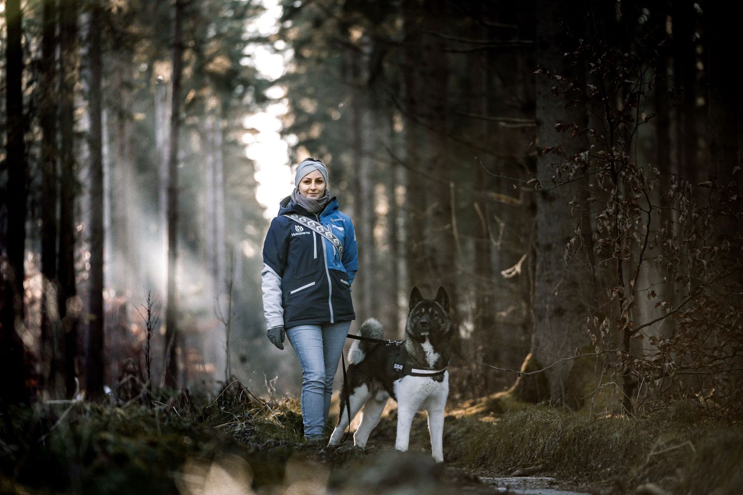 A person walks their dog along a misty forest trail as sunbeams stream through the trees.