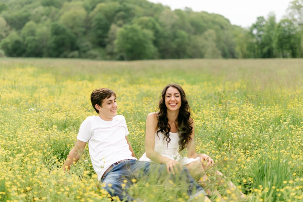 A smiling couple sits together in a sunny field of yellow wildflowers.