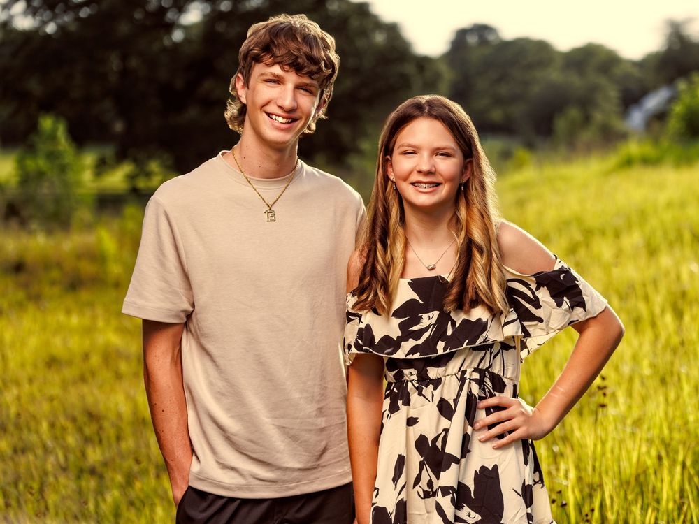 Two smiling teenagers pose together in a sunny green field, wearing casual summer outfits.