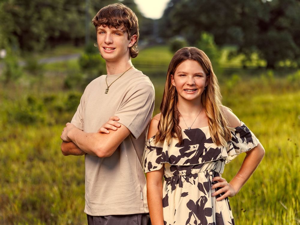 Two smiling young siblings posing outdoors in a green field, boy in tan shirt and girl in floral top.