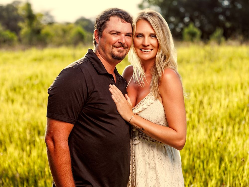 Smiling couple posing together in a lush green field, man in black polo and woman in white dress.