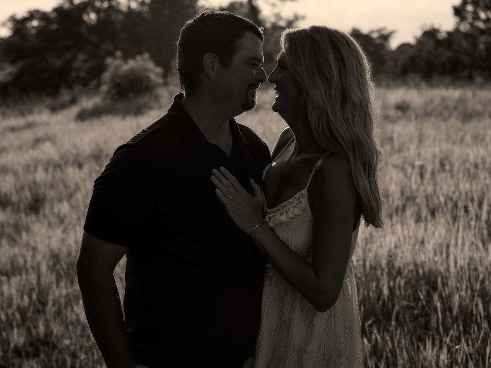 Black and white photo of a romantic couple gazing at each other in a grassy field at dusk.