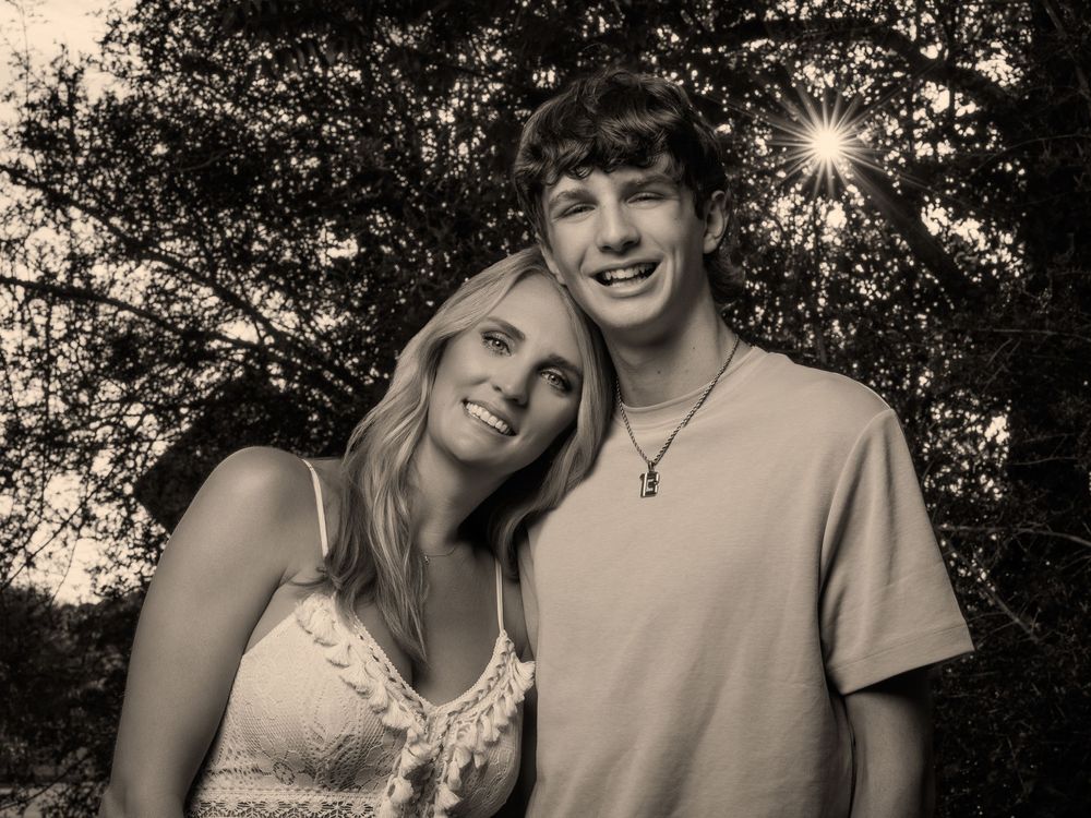 Black and white photo of a smiling young couple posing together outdoors among trees in warm sunlight.