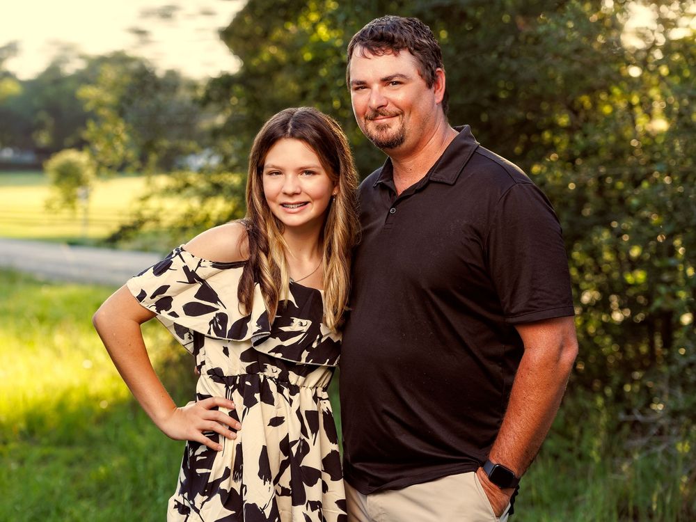 A smiling couple poses outdoors near lush green trees, woman in floral dress and man in black polo shirt.