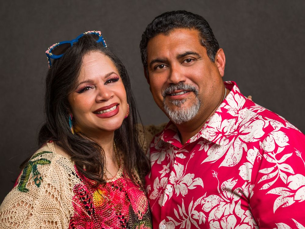 A couple wearing colorful Hawaiian-style clothing smile together in a studio portrait.