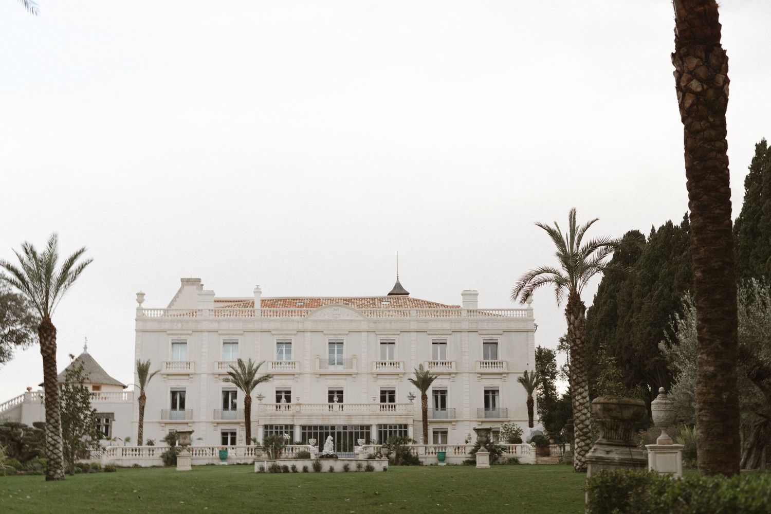 A grand white Mediterranean-style villa with palm trees and manicured grounds under an overcast sky.