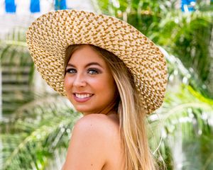 Smiling young blonde woman wearing large straw sun hat with tropical palm trees background.