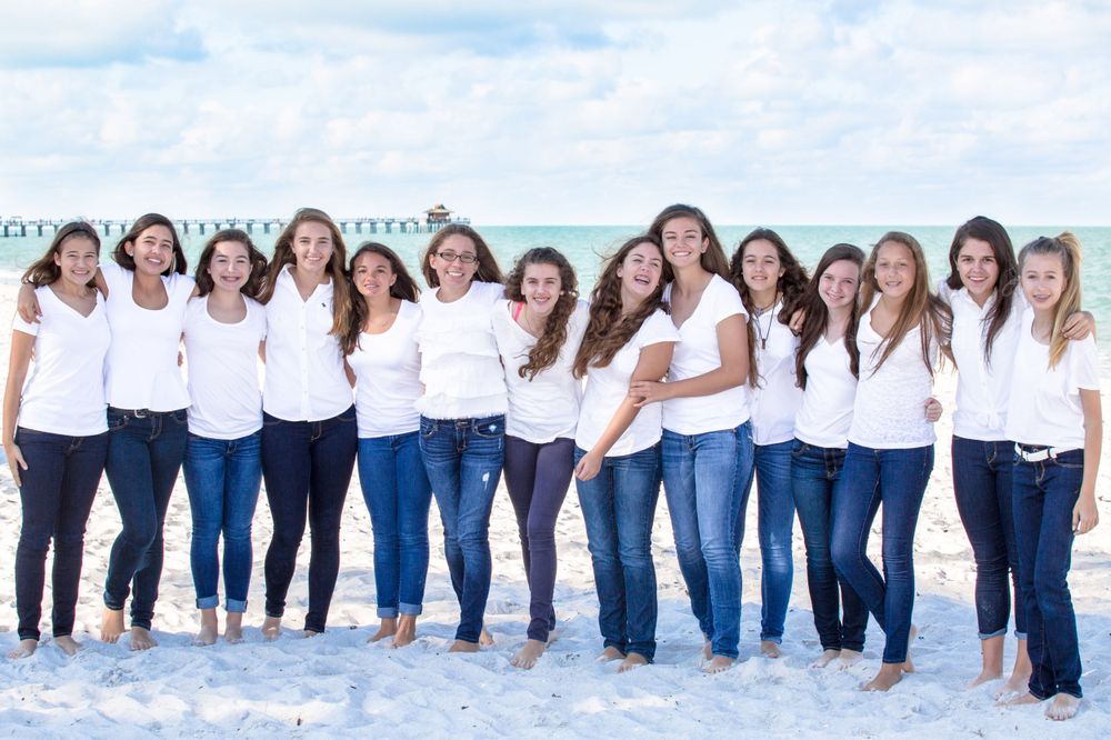 Group of teenage girls in matching white tops and jeans posing together on a beach.