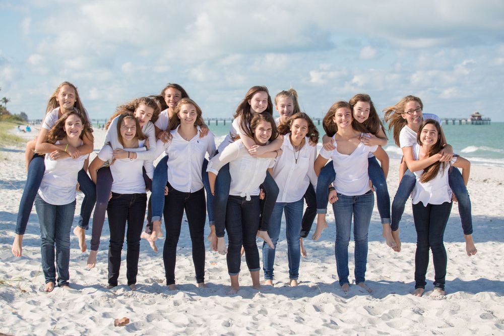 Group of teenage girls in white shirts and jeans doing piggyback rides on a sunny beach.
