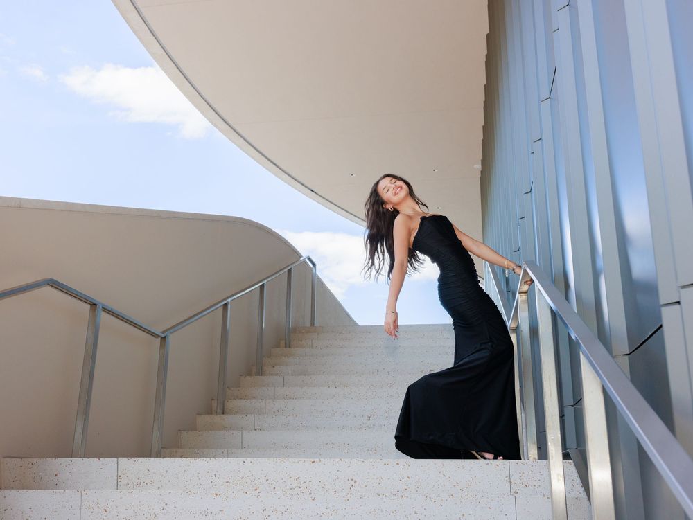 Woman in elegant black gown posing gracefully on white curved staircase with modern architecture and blue sky.