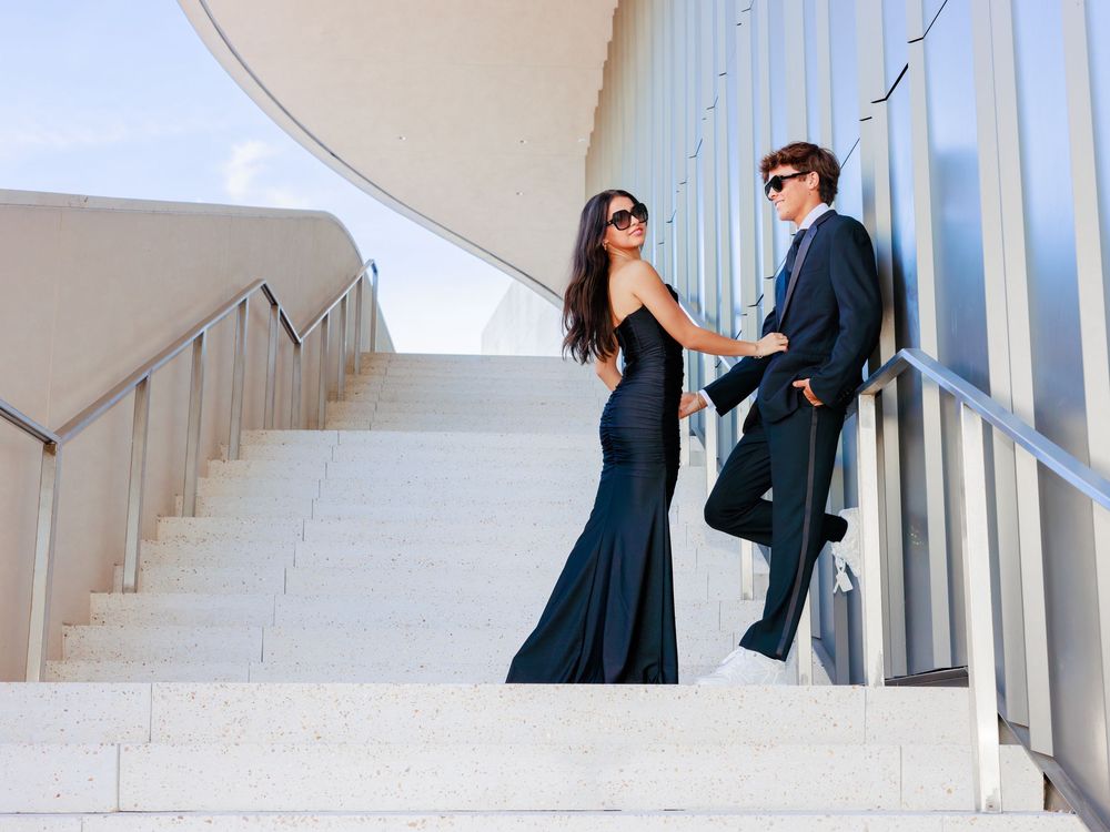 Elegant couple in formal attire posing on white marble stairs, woman in black gown, man in dark suit.