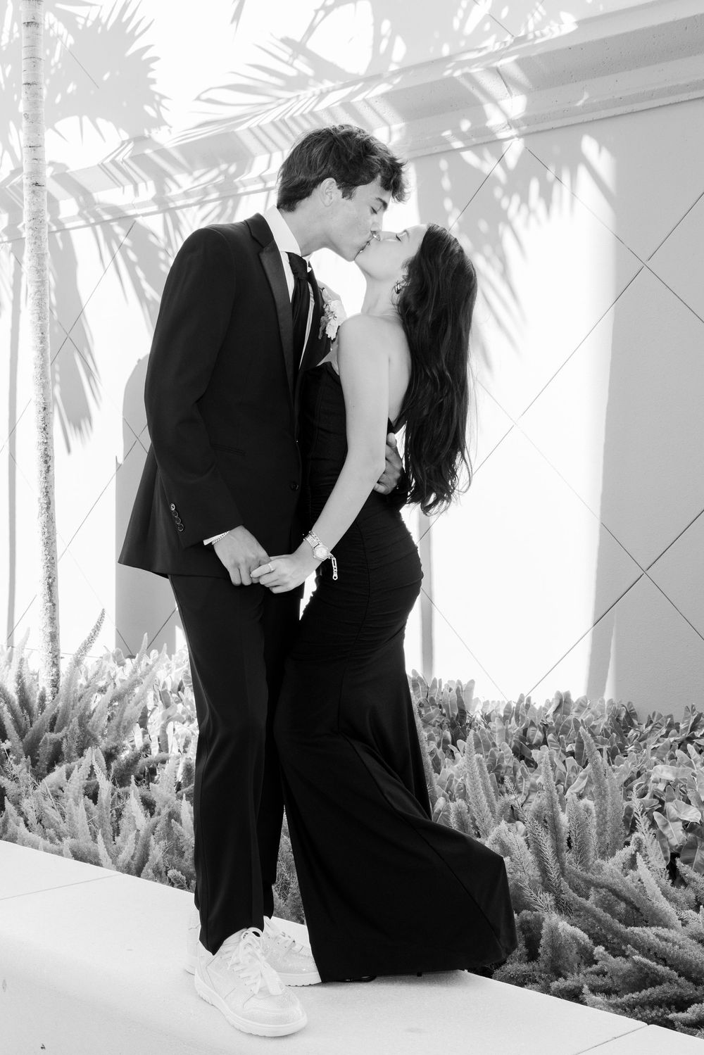 Black and white photo of a couple kissing, man in tuxedo and woman in elegant black gown, surrounded by tropical plants.