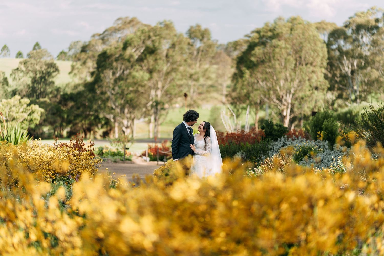 Couple standing in a field of golden wildflowers with trees in the background.