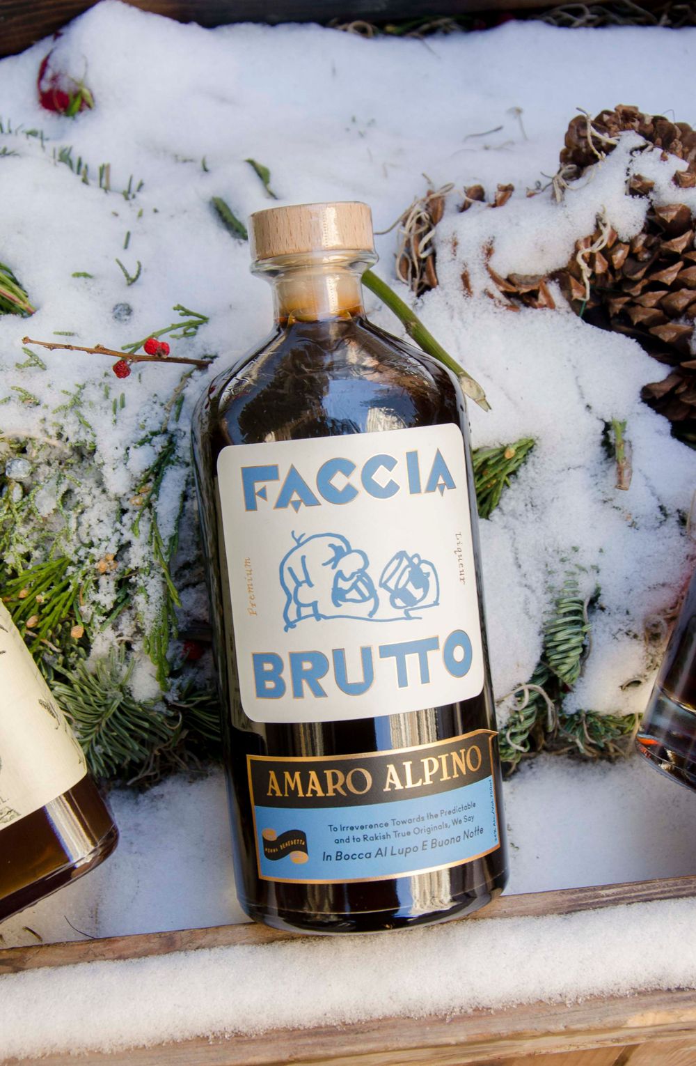 Bottle of Faccia Bruto Amaro Alpino liqueur photographed in snow with pine cones.