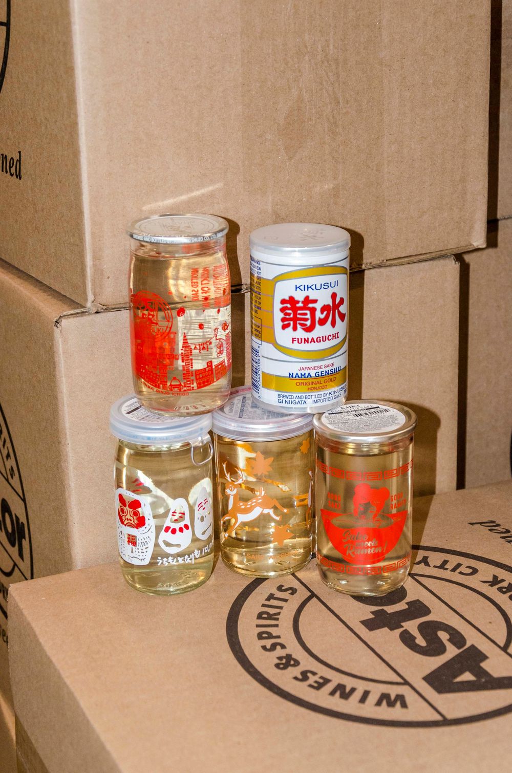 Several cans and jars of Japanese sake and alcoholic beverages displayed on brown cardboard boxes with visible branding.